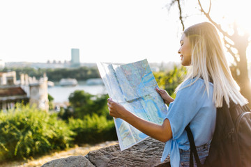 Wall Mural - Young female traveler standing in front of beautiful city view and looking at the map 