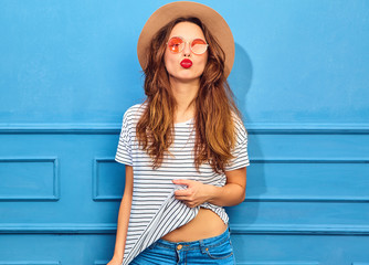 Young stylish girl model in casual summer clothes and brown hat with red lips, posing near blue wall. Giving air kiss