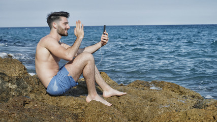 Young shirtless muscular handsome man doing videochat with smartphone by the sea with earphones