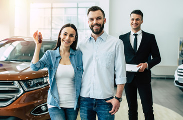 Wall Mural - We bought a car. Young and smiling man and woman show the keys to a new jeep looking at the camera on the background of the dealership