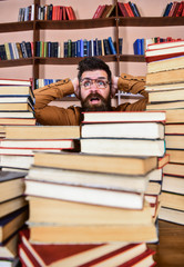 Leinwandbilder - Teacher or student with beard wears eyeglasses, sits at table with books, defocused. Man on shocked face between piles of books in library, bookshelves on background. Scientific discovery concept.