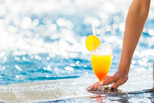 Womans Manicured Hand Touching A Cocktail With An Orange On A Glass Standing On The Edge Of The Pool. Free Space For Advertising Text