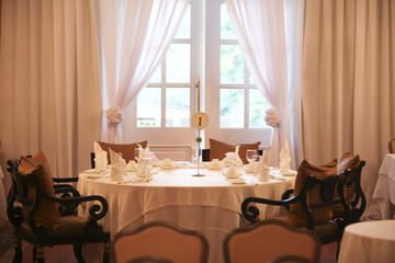 Close-up of a big round numbered table in front of a French window in a festive colonial dining room with white sheer curtains, a white tablecloth and white napkins.