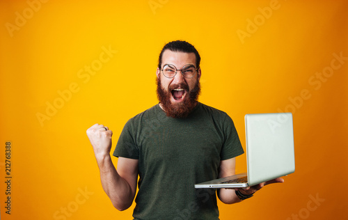 Fotografiet Winner bearded man wearing glasses and looking at the camera holding his notebook and making a winner gesture