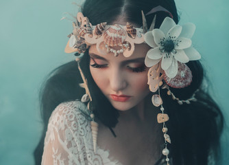 Close-up portrait - makeup of a river nymph, in gentle green, yellow, peach hues. The brunette girl with an unusual wreath with seashells. Puppet's baby face. Background thick fog.