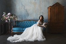 Sexy And Beautiful Brunette Model Girl In Stylish And Fashionable Lace Wedding Dress With Naked Shoulders Sits On The Antique Sofa And Posing In Luxury Vintage Interior