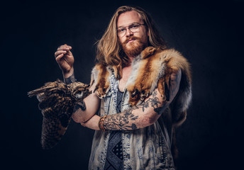 Wall Mural - Portrait of a tattoed redhead hipster male with long luxuriant hair and full beard dressed in a t-shirt and jacket holds a keeps the scarecrow of an owl and fox skin on a shoulder in a studio.