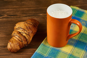 Chocolate croissant on a yellow-blue napkin and orange mug with milk on a brown wooden background