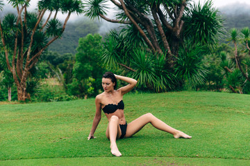 Wall Mural - A young European-looking woman with a tanned body in a black bathing suit relaxes sitting on the green grass, straightens her hair with her hand, relaxes in the rain.