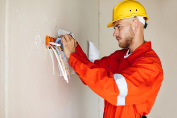 Wall Mural - Young builder in orange work clothes and yellow hardhat thoughtfully working with wires while doing repair of flat