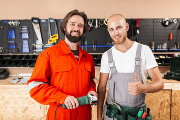 Wall Mural - Two smiling builders in work clothes happily looking in camera spending time in workshop with tools on background