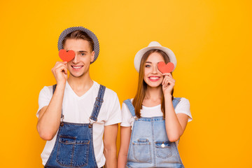 Young happy students couple in love wearing hats holding two little red paper hearts on eyes, isolated