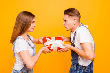 Portrait of lovely young nice cute couple seriously competing for red present box with white bow, ribbon, isolated over yellow background