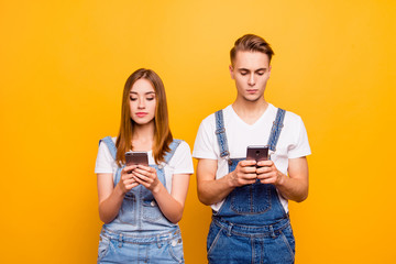 Wall Mural - Portrait of cute young couple wearing casual, holding their smartphones in hands, seriously looking at screens, over yellow background, isolated