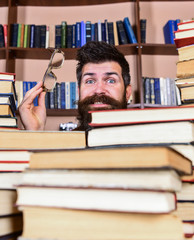 Leinwandbilder - Teacher or student with beard wears eyeglasses, sits at table with books, defocused. Man on surprised face between piles of books in library, bookshelves on background. Scientific research concept.