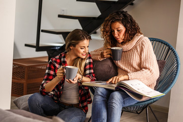 Two best female friends looking at family album.They sitting in living room and drinks coffee.