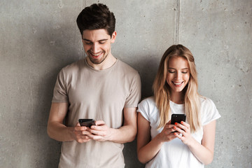 Poster - Photo of positive people man and woman smiling while both using mobile phones, isolated over concrete gray wall indoor