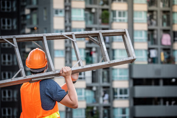 Wall Mural - Young Asian maintenance worker with orange safety helmet and vest carrying aluminium step ladder at construction site. Civil engineering, Architecture builder and building service concepts