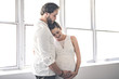 © Louis-Photo - Handsome man and his beautiful pregnant wife standing near the window at home