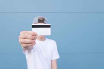 Wall Mural - Young man in a white T-shirt with a bank card his hand stands on a blue pastel background. Man shows a credit card in a camera. Bank card in hand close-up focus