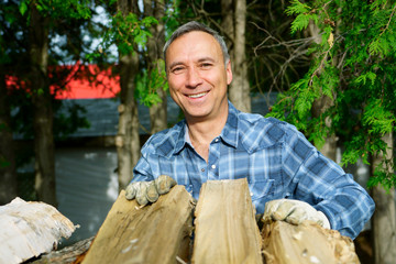 A 50 years old caucasian man wearing a bleu shirt is smiling at us while piling his wood for the winter season, in order to heat the house.