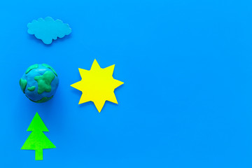 Wall Mural - Environment protection concept. Plastiline symbol of planet Earth and sun, cloud, tree coutout on blue background top view copy space