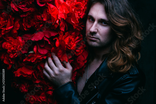 Fotografía brutal man in a leather jacket on a naked body with long hair stands near the decor with red flowers flowers