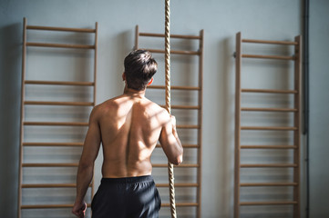 Wall Mural - Fit young man in gym standing topless , holding a climbing rope. Rear view.