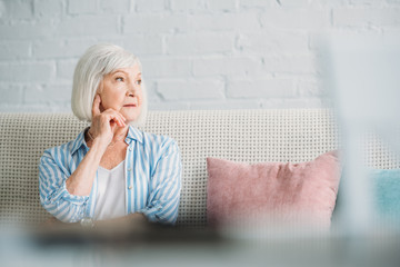 portrait of pensive grey hair woman looking away resting on sofa at home