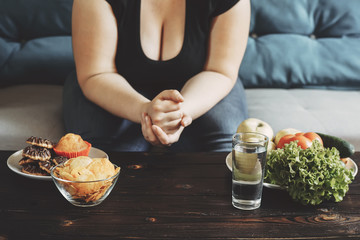 Wall Mural - Young overweight woman bored of diets choosing between healthy and junk food. Dieting, healthy food, weight losing, slimming