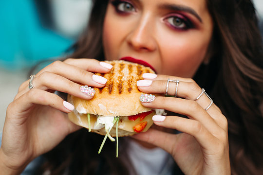 Close-up of unfocused girl with bright make up holding delicious crunchy cheeseburger in hands with perfect nude manicure, wearing rings.