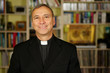 A good looking catholic priest is studying into his library. He looks at us with serenity, peace and optimism.