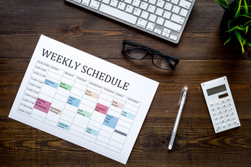 Weekly schedule of manager, office worker, pr specialist or marketing expert. Table with multicolored blocks on dark wooden office desk with computer, glasses, calculator top view