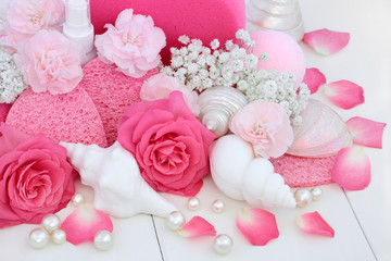 Wall Mural - Cleansing spa beauty treatment products with pink roses and carnation flowers, shell shaped soaps, bath bomb, sponges with decorative seashells and pearls on white wood background.