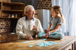 Joyful aged man assembling jigsaw puzzles with his granddaughter