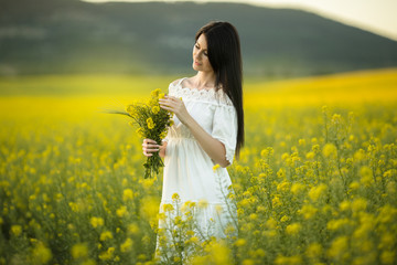 Happy young woman with bouquet of wildflowers in yellow field in sunset lights, summer time