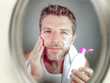 © TheVisualsYouNeed - young happy and attractive man applying moisturizer lotion or anti aging beauty cream smiling confident and conscious looking at bathroom mirror