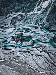 A drone shot from above on a green and blue metallic looking glacier river stream in Iceland with repeating grey, beige and dark green patterns, textures and structures running from top to down