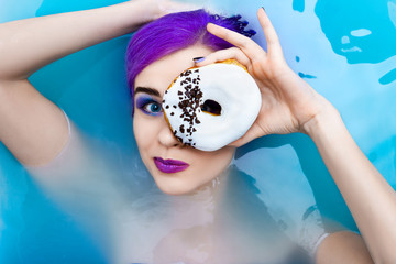 Portrait of young style girl female adult  woman / fashion luxury model with purple hair relaxing in bathtub with sweet donut isolated by blue water. Organic skin care, beauty and body care concept.