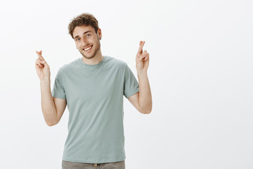 Carefree charming european man in earrings with blond hair, tilting head and smiling with friendly expression, raising crossed fingers, staying positive while waiting for results and wishing