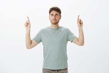 Wall Mural - Casual good-looking male coworker in t-shirt, raising index fingers and pointing up while making indifferent annoyed grimace, being fed up with noise colleague and showing direction office upstairs