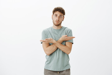 Poster - Studio shot of confused and troubled handsome male coworker in trendy t-shirt, crossing hands over chest and pointing left and right, being questioned what direction choose, standing over gray wall