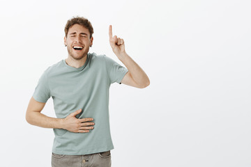Wall Mural - Guy likes woman with sense of humour. Portrait of handsome funny male model in casual t-shirt, laughing out loud with closed eyes and broad smile, pointing up and holding hand on belly