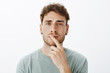© Cookie Studio - Portrait of smart focused attractive guy in earrings with short curly hair, holding index finger on lip and gazing with thoughtful expression at camera, thinking or making decision