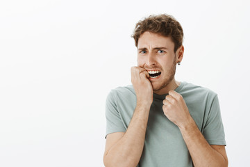 Wall Mural - Portrait of stressed anxious european male with fair hair, biting fingernail and frowning, grimacing from fright, being nervous and afraid, standing shocked over gray background