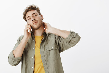 Wall Mural - Confident hot curly-haired male model with beard, lifting head and touching neck while talking on smartphone, standing self-assured and flirty over gray background, gazing with sensual look