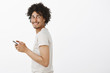 Studio shot of carefree charming and happy male with afro haircut, standing in profile, turning backwards and smiling joyfully, holding smartphone, being interrupted from playin app