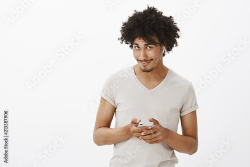 Good Looking Flirty And Joyful Hispanic Guy With Moustache And Curly Hair Looking From Under Forehead With Cute Sensual Smile Holding Smartphone And Wearing Wireless Earphones Using App Stock Photo Adobe Stock