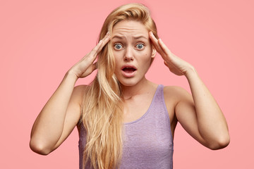 Wall Mural - Stressed young woman keeps hands on temples, notices or remembers something terrific, has scared expression, dressed casually, stands indoor. Beautiful blonde female looks nervously at camera
