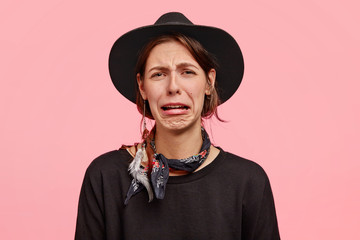 Wall Mural - Photo of sorrowful female cries as has grief, purses lips and has discontent facial expression, wears elegant black hat and sweater, poses against pink background. Negative emotions, feelings concept